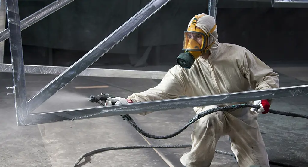 Arbeiter in Schutzkleidung, der eine große Metallstruktur mit Spritzlackierung beschichtet.